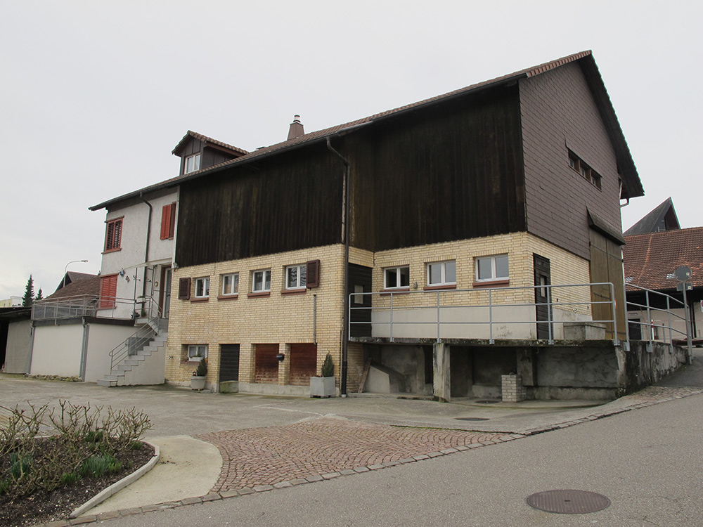 Das Bauernhaus oben wurde 1940 mitten in Dielsdorf gebaut – zu einer Zeit, als Bauernhöfe fast nur noch ausserhalb der Dörfer errichtet wurden. Es ersetzte einen abgebrannten Vorgängerbau. Das in einer gemässigt modernen Formensprache gestaltete Haus erinnert als Vielzweckbau mit Wohn- und Ökonomieteil unter einem Dach zwar an das traditionelle Bauernhaus. Stall und Scheune sind jedoch nicht wie üblich quer, sondern längs zum First ausgerichtet und die Eingänge stirnseitig angelegt. Auch öffnen sich die Fenster des Stalls gegen Süden und lassen viel Licht ins Innere, womit sie ein Postulat der Moderne nach mehr Licht und Luft umsetzen. (Bild: zVg)